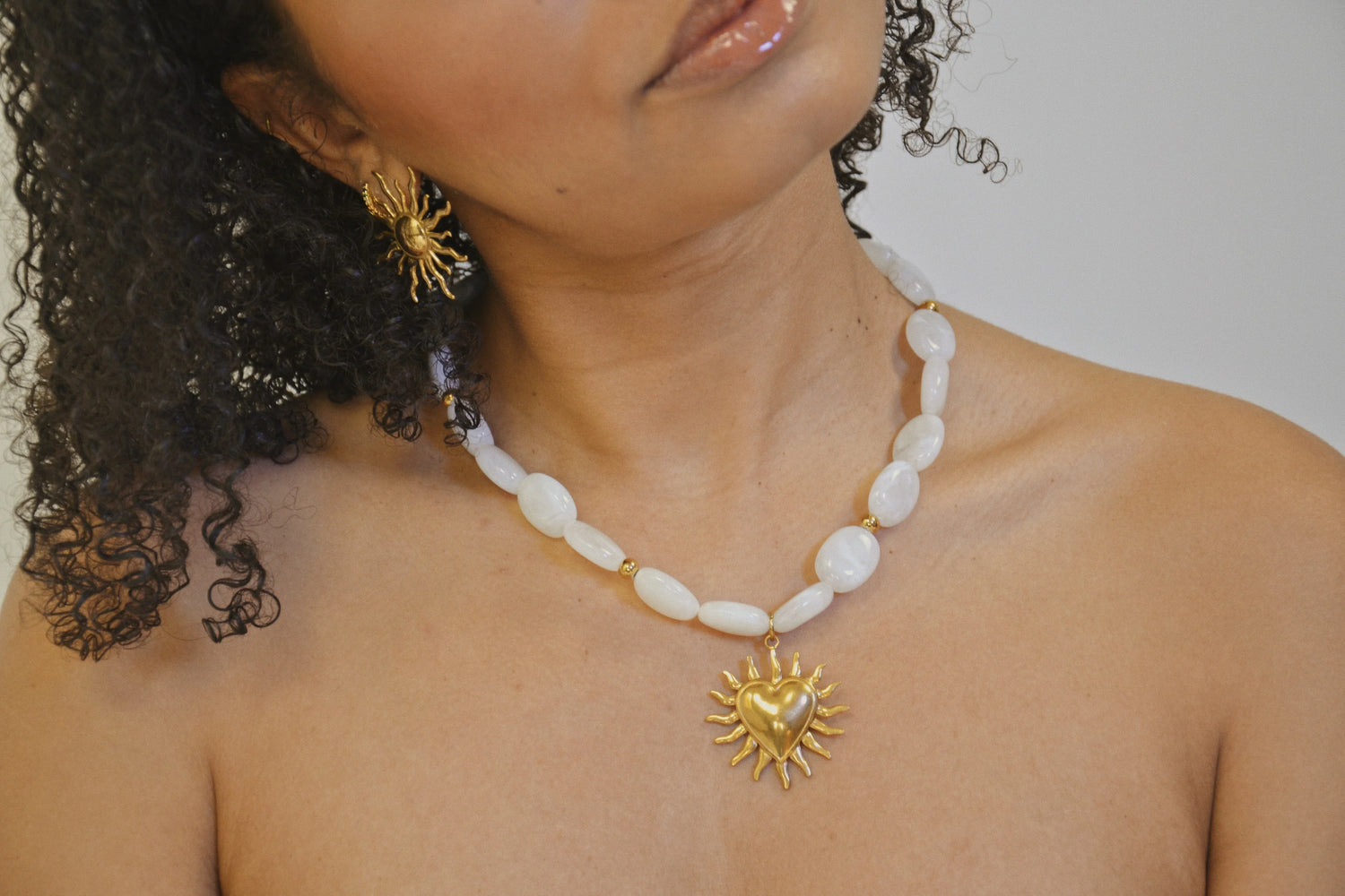 Femme portant un collier de perles blanches avec pendentif cœur soleil doré et boucle d’oreille assortie