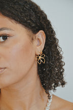 Femme portant une boucle d’oreille fleur dorée et collier perles, fond neutre